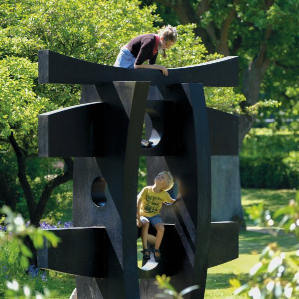 Two children play on the large black sculpture ‘Pagoda’ by Hans August Andersen in the ‘Geografisk Have’. The sculpture is surrounded by lush nature and green trees. 